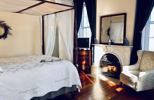 A queen canopy bed with a white bed spread facing a marble fireplace and an upholstered chair/
